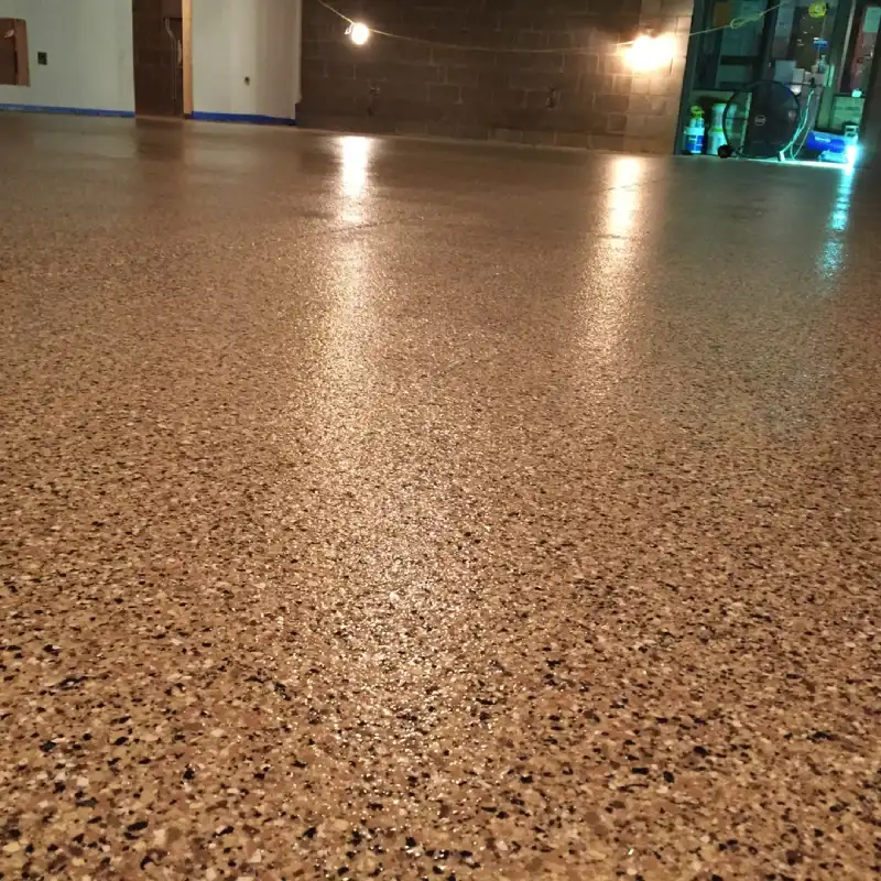 A glossy, newly finished chip epoxy flooring in a garage or workshop. The floor is covered in a decorative mix of beige, brown, and black vinyl flakes